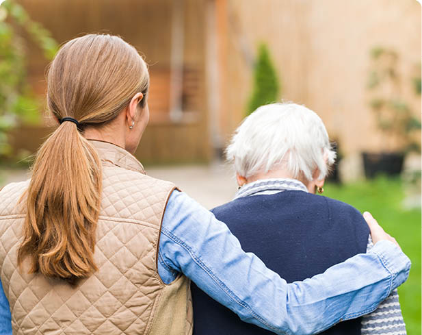 One person with long hair in a ponytail wearing a quilted vest and denim shirt has their arm around another person with short white hair wearing a dark sweater, both seen from behind in an outdoor setting. Description generated by AI