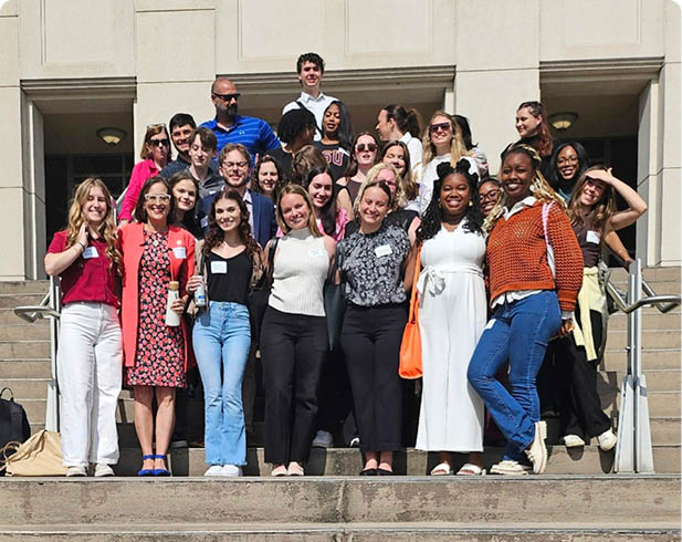 A group of adults dressed in casual and semi-formal clothing are standing on outdoor steps, smiling and posing for a group photo in front of a building. Description generated by AI