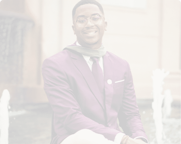 A person wearing a purple blazer, beige pants, and a tie is sitting cross-legged near a water fountain. Description generated by AI