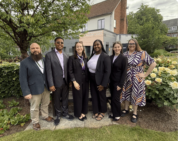 Six adults dressed in business and casual attire stand together outdoors in front of a building with greenery and flowers around them. Description generated by AI