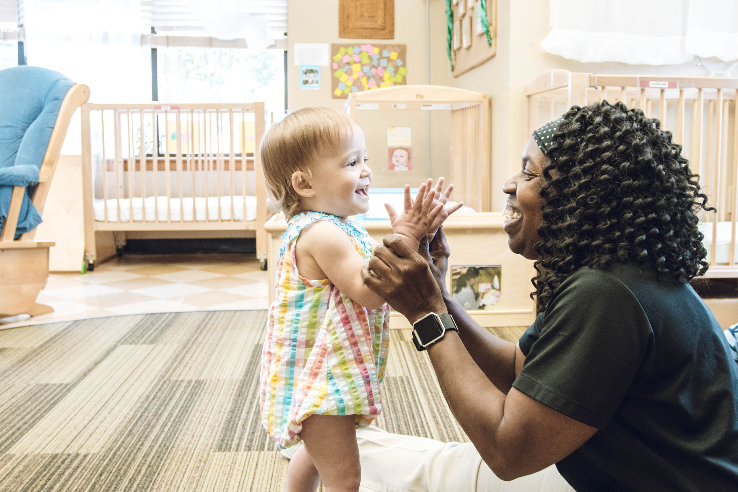 female toddler smiling with Black female teacher holding her as she stands and smiling