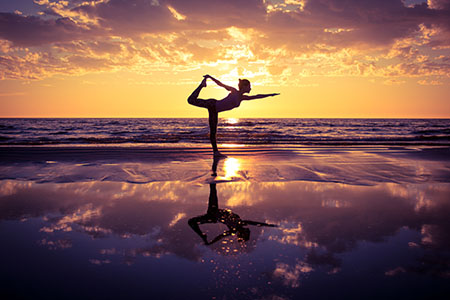Silhoutte of a woman on a beach at sunrise in standing bow pose.