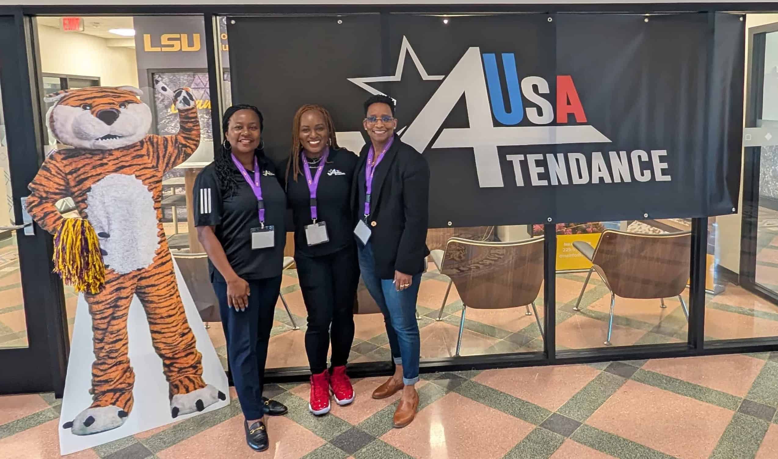 Three women standing next to Mike the Tiger and in front of the Attendance USA flag in Huey P. Long Field House
