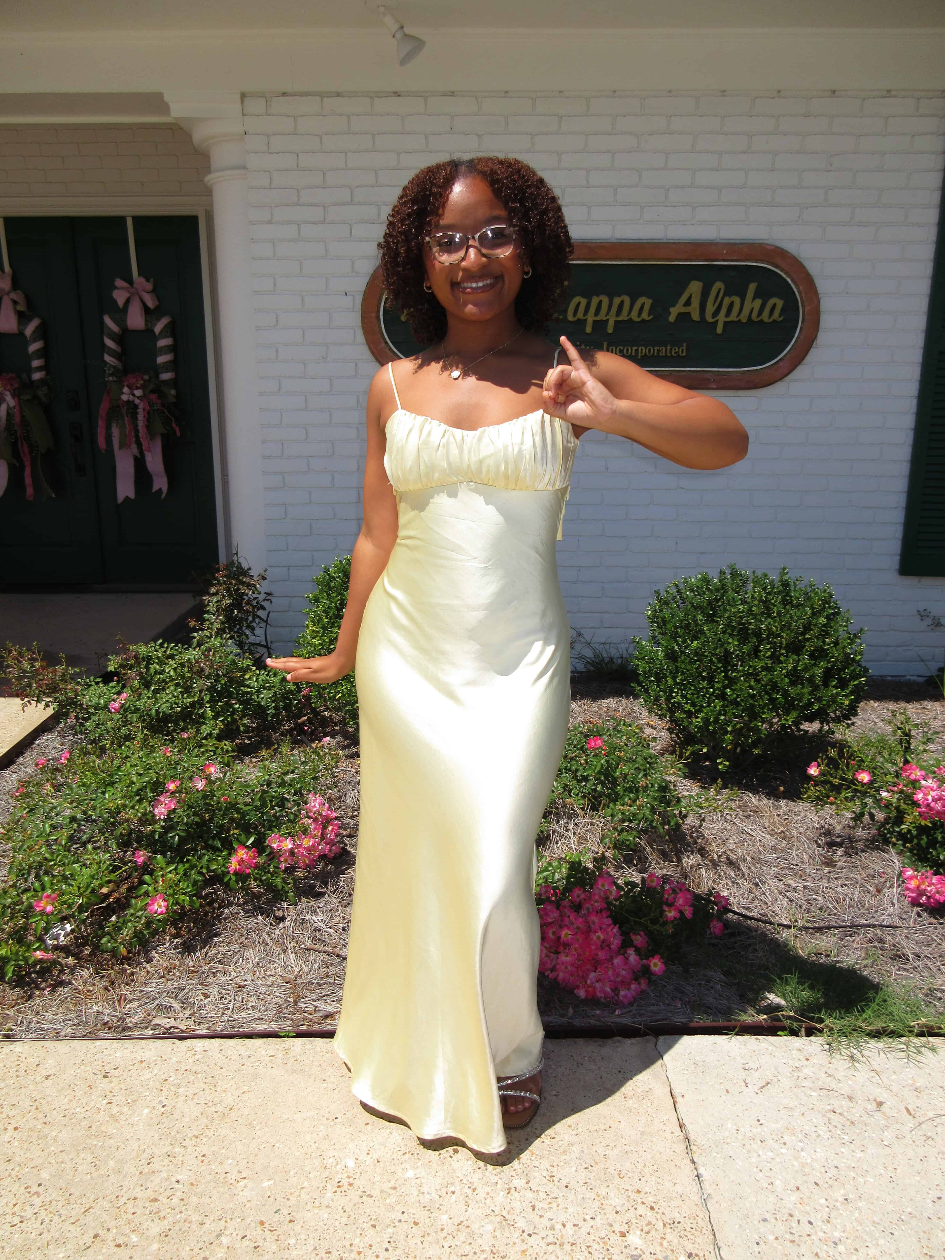 Paris Bickham standing in a yellow gown holding up her pinky finger representing the Alpha Kappa Alpha sorority