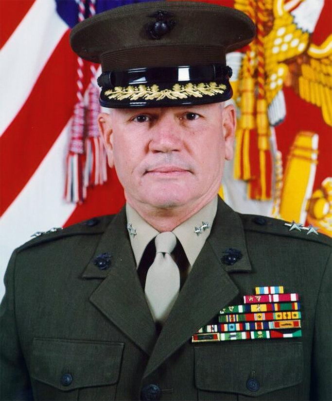 Official portrait of Maj. Gen. Ron Richard in his Marine Corps service uniform and cap, with two-star rank insignia and rows of ribbons, in front of the U.S. and Marine Corps flags.