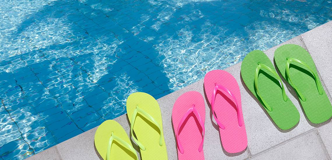 Brightly colored flip-flops lay on a pool deck next to bright blue water.