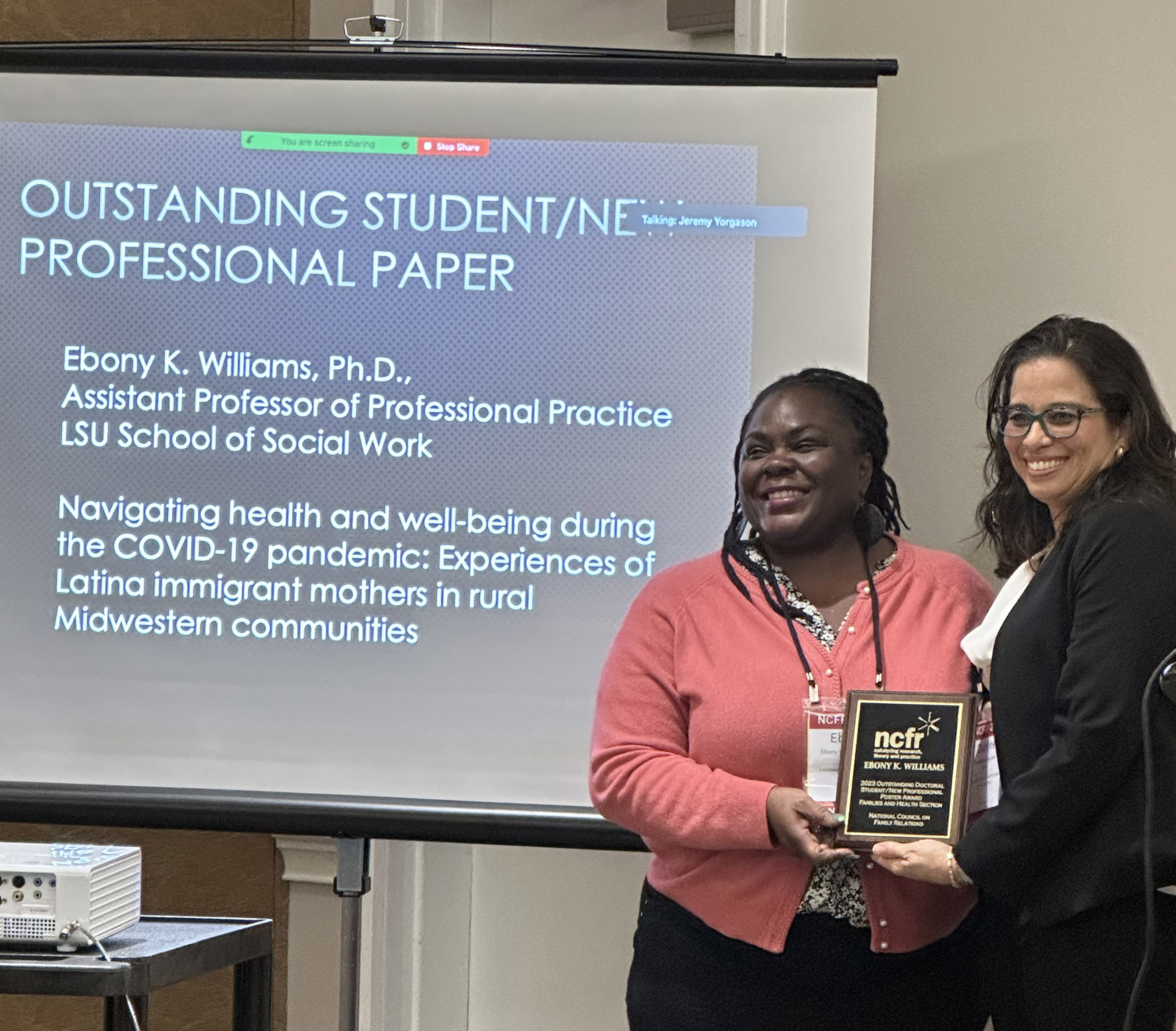 Dr. Ebony Williams receives award plaque from Daphne Hernandez in front of a description of the award
