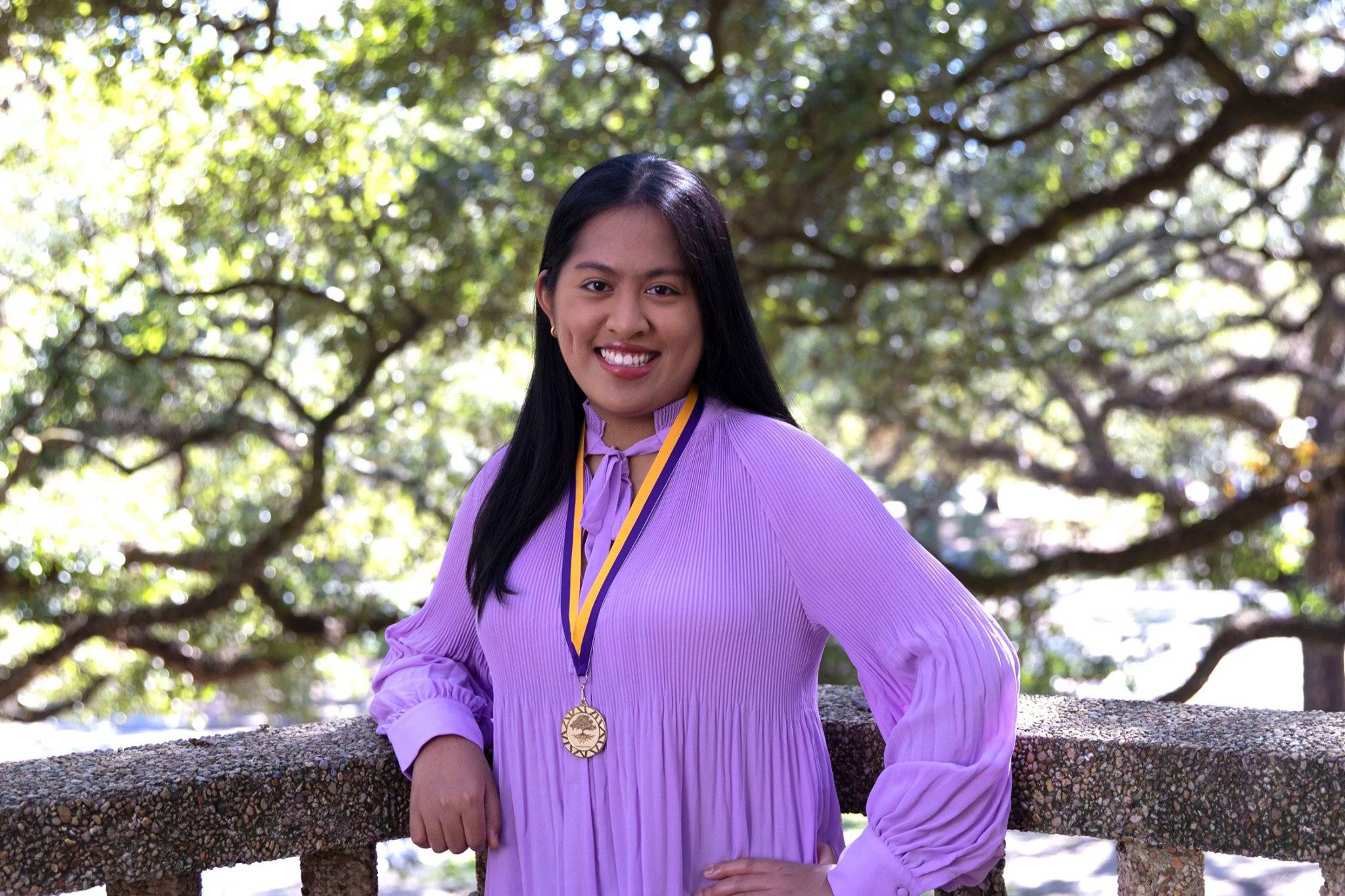 Mae Anne Mangaoil from the waist up, smiling, wearing a long-sleeved purple shirt and a medal with purple and gold ribbon around her neck, outdoors in front of a tree