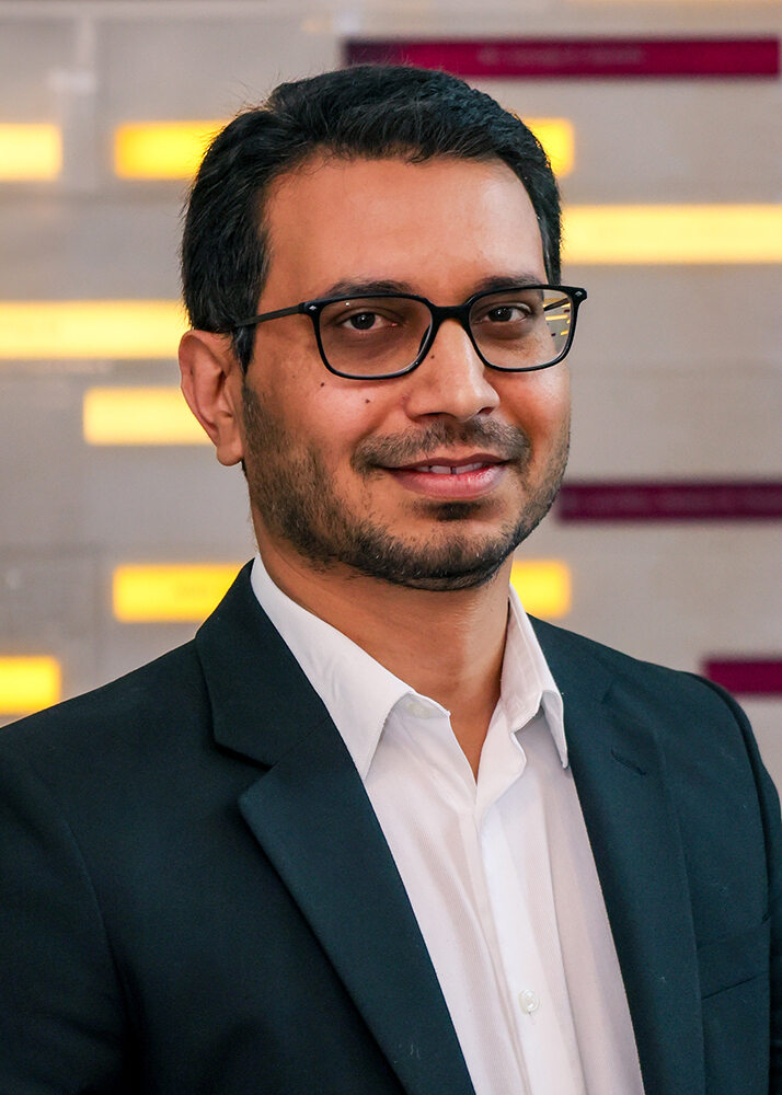 Portrait headshot of Dr. Umar Farooq in professional dress clothes in front of a soft-focused background of colorful streaked lights.