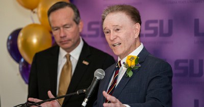Roger Ogden speaking at a podium with LSU President F. King Alexander in the background