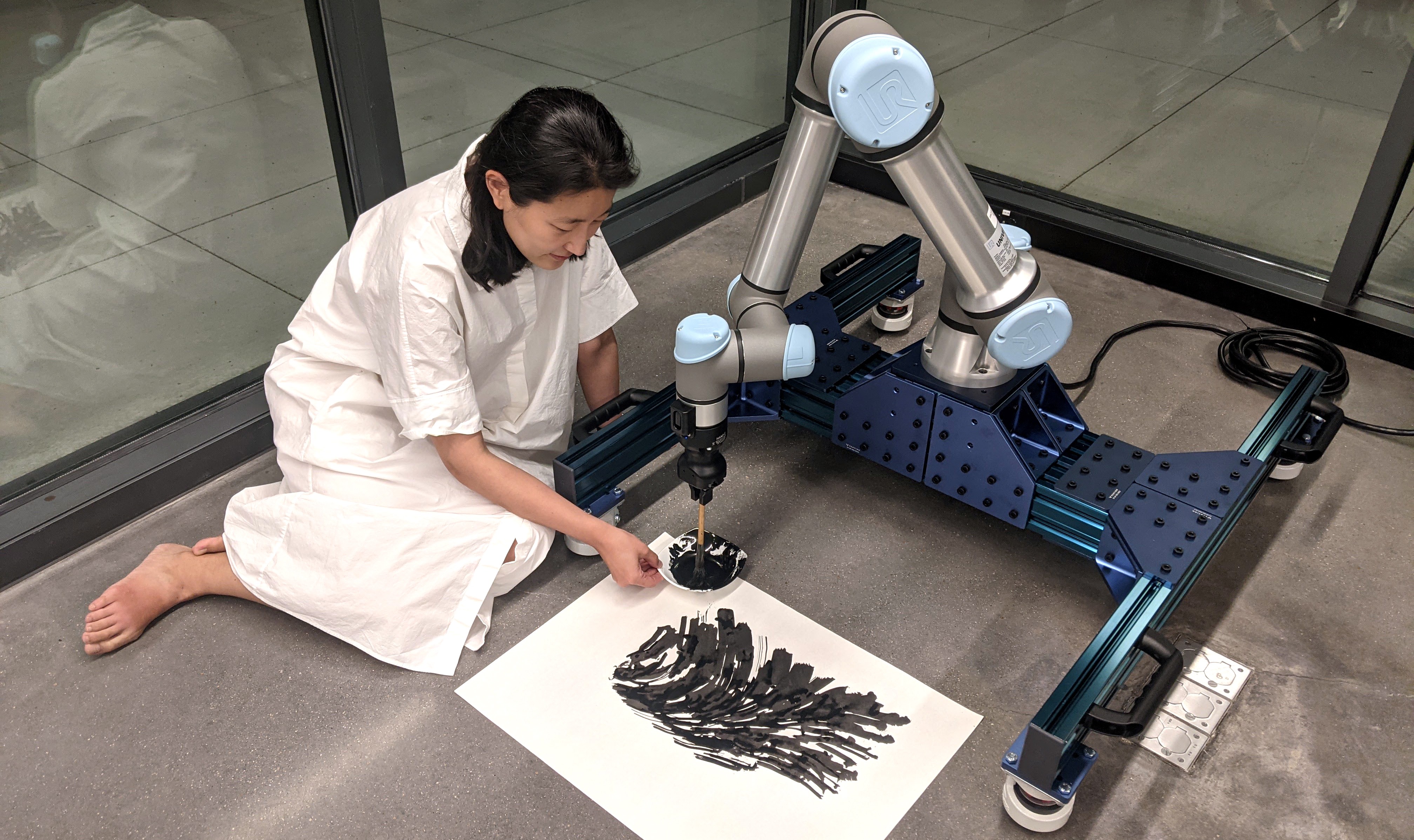 A robot with a paintbrush with a woman sitting on the floor holding a bowl of paint