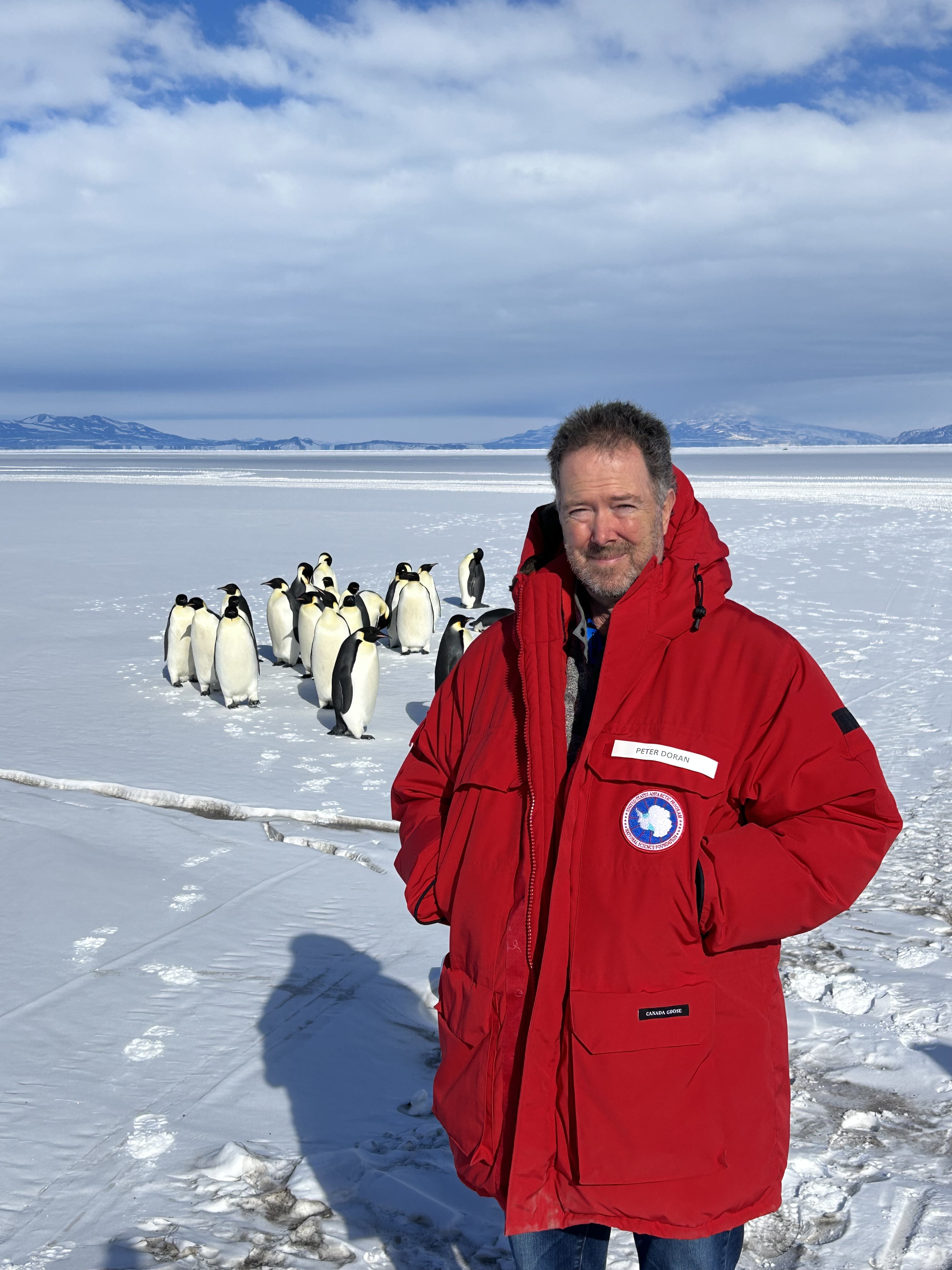 Peter Doran in Antarctica with his team of Emperor Penguins. OR Peter Doran in front of McMurdo Station, Antarctica in 2025