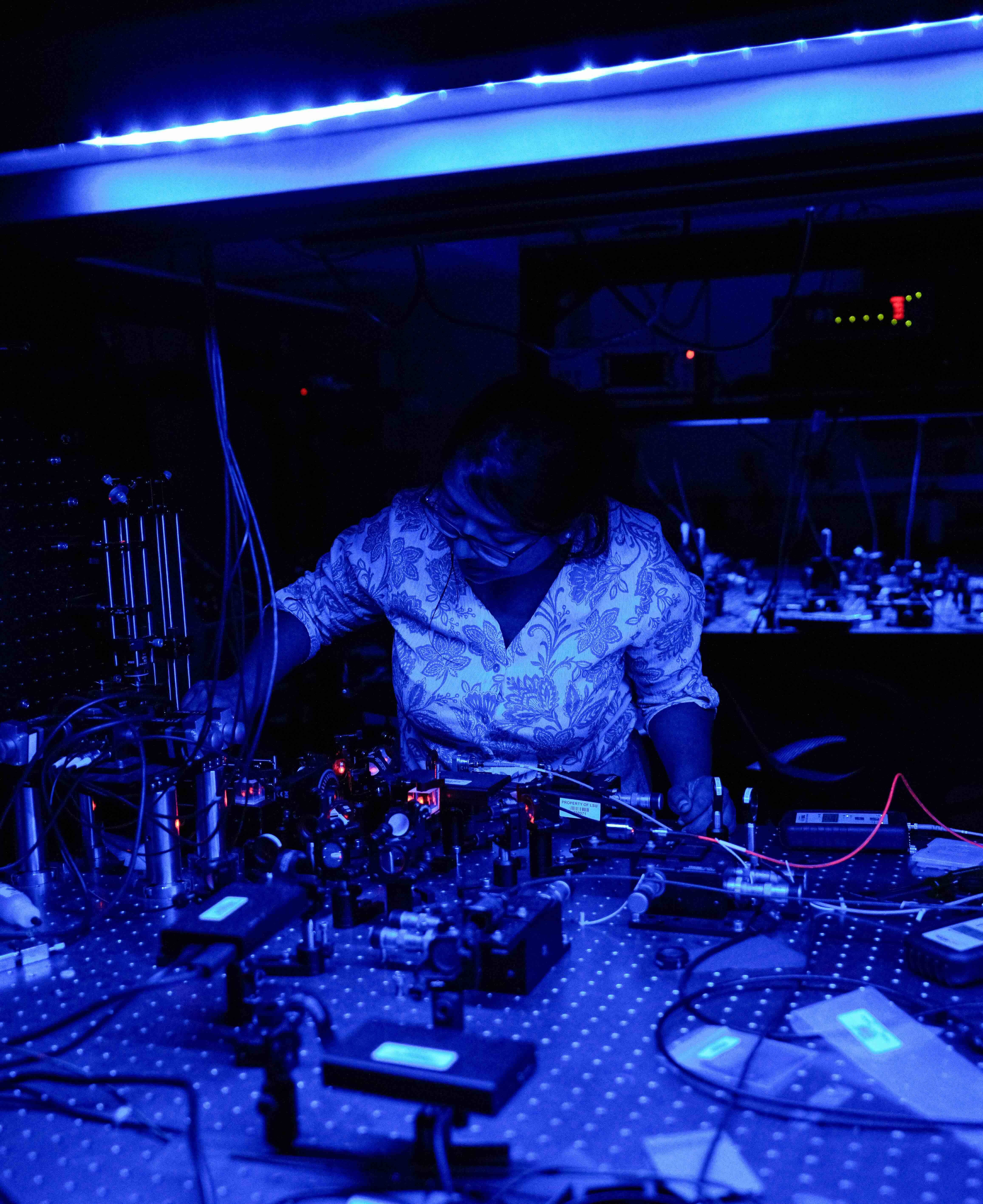 Graduate researcher Rima Bhandari adjusts a laser-based quantum optics experiment in a dark laboratory illuminated by blue light.