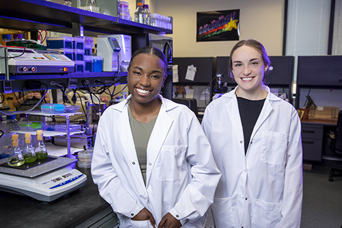 Dykia Williams and Lexie Cheramie, adorned in lab coats, share smiles while positioned in the Vinyard Lab at Louisiana State University. Their cheerful demeanor reflects the positive and collaborative environment within the laboratory setting.