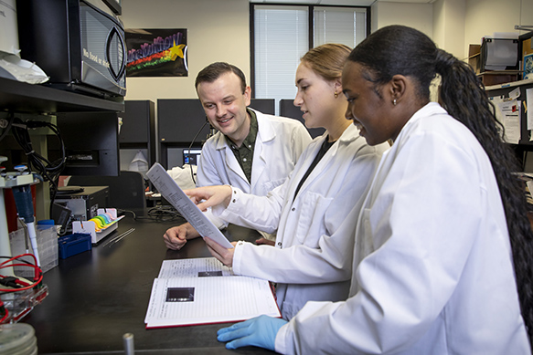 Beckman Scholars Dykia Williams and Lexi Cheramie engaged in a discussion about scientific findings, seated with Professor David Vinyard in the Vinyard Lab at Louisiana State University. The collaborative atmosphere reflects their commitment to academic inquiry and research excellence.