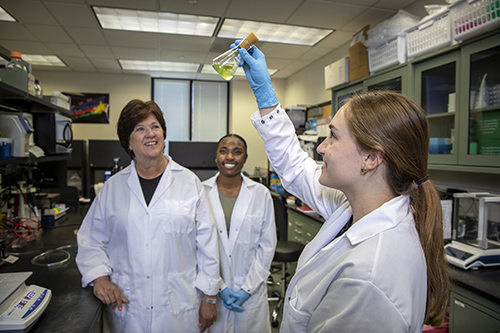 Beckman Scholar Lexi Cheramie carefully holds a corked Erlenmeyer flask containing a vibrant neon green liquid, with her colleague Dykia Williams and College of Science Dean Cynthia Peterson smiling in the background. They are actively involved in collaborative laboratory work at Louisiana State University.