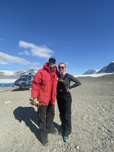 Senior geophysics major Leora Wilson and Dr. Peter Doran in Antarctica, with snow-capped mountains and a helicopter in the background near Lake Fryxell in the McMurdo Dry Valleys of East Antarctica.