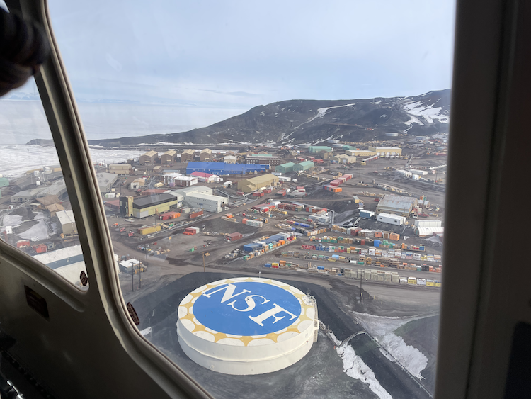 National Science Foundation logistics hub near the landing strip, captured from Leora Wilson's helicopter, providing a perspective of McMurdo Station, the largest settlement in Antarctica, accommodating up to 1,000 people during the summer months.