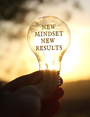 Photo of a hand holding a lightbulb in front of a sunrise. The lightbulb contains the words New Mindset New Results.